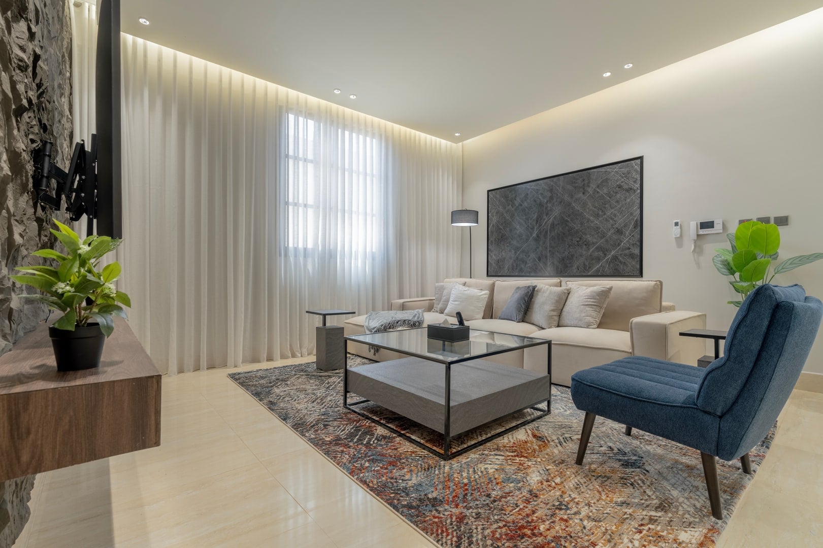 Living room with a beige sofa, a glass coffee table, a blue armchair and a large framed marble print, Riyadh apartment