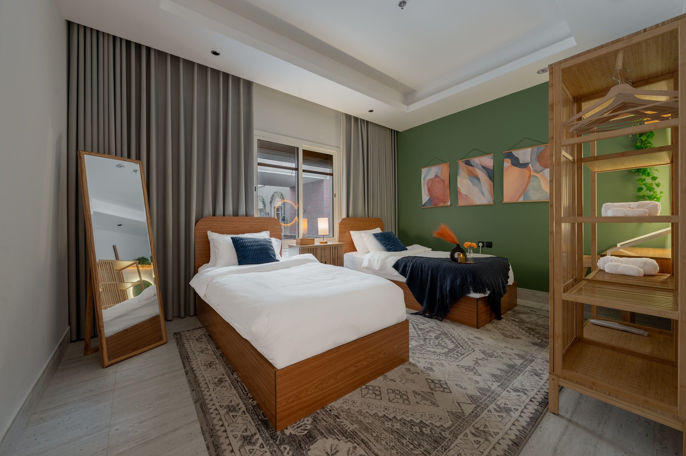 Bedroom with two single wood beds, three prints on a green wall, a floor mirror and an open bamboo wardrobe, Riyadh apartment