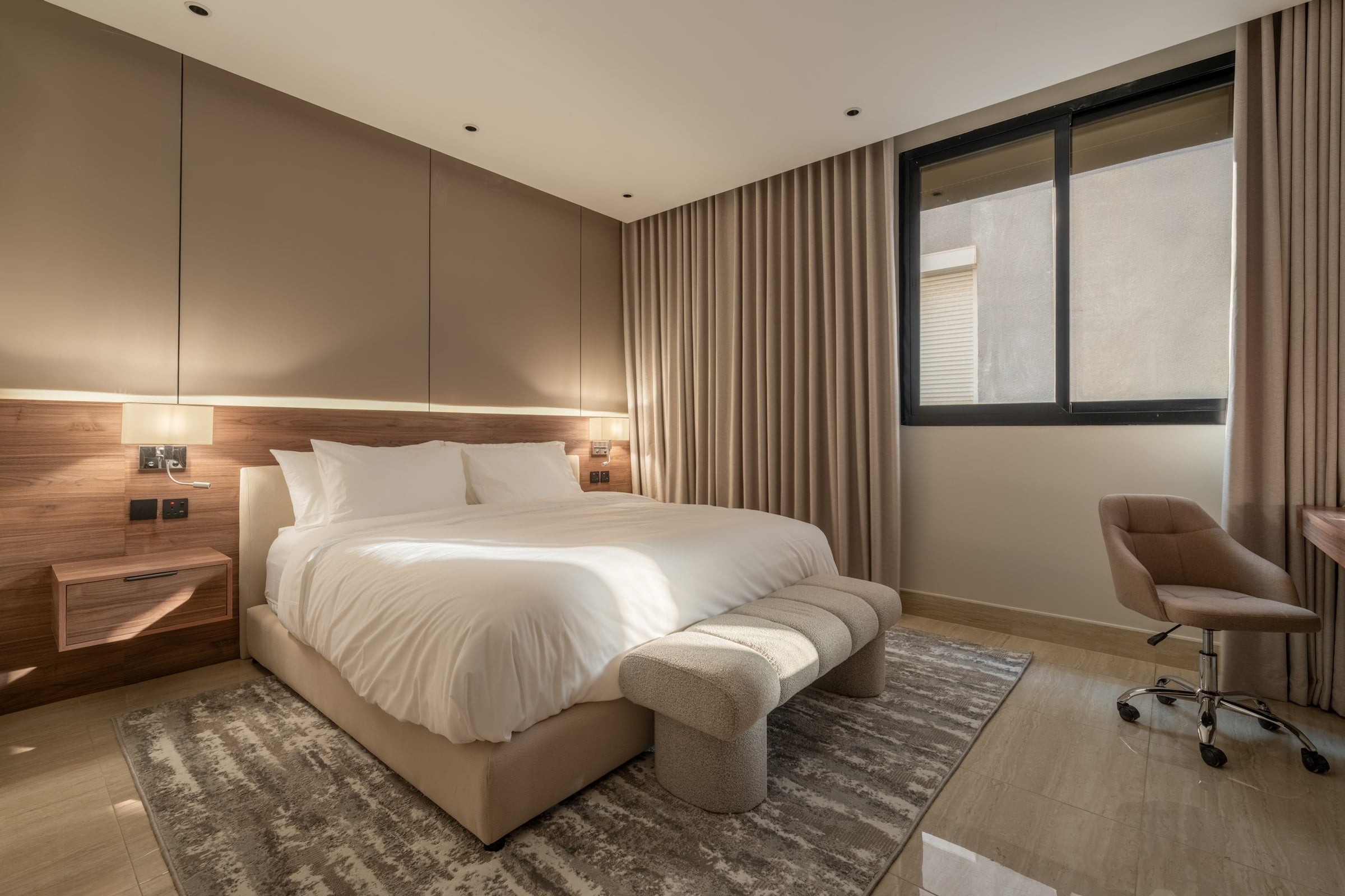 Bedroom with a double bed, a walnut headboard wall, an upholstered bench and a desk chair, Riyadh apartment