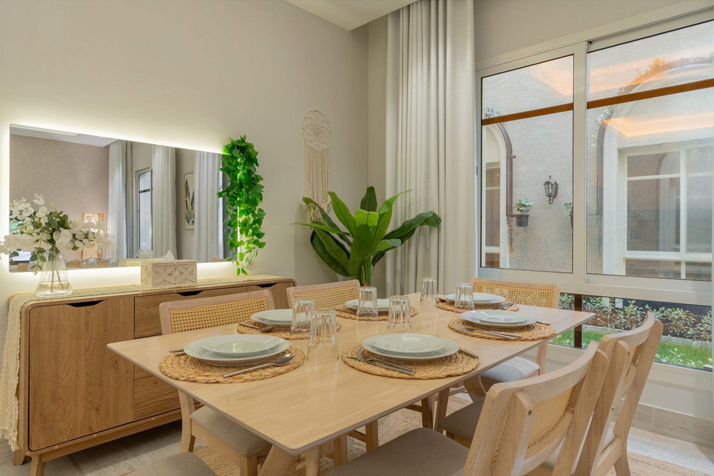 Light wood dining table for six with cane-back chairs, a sideboard and a backlit mirror beside a garden window, Riyadh apartment